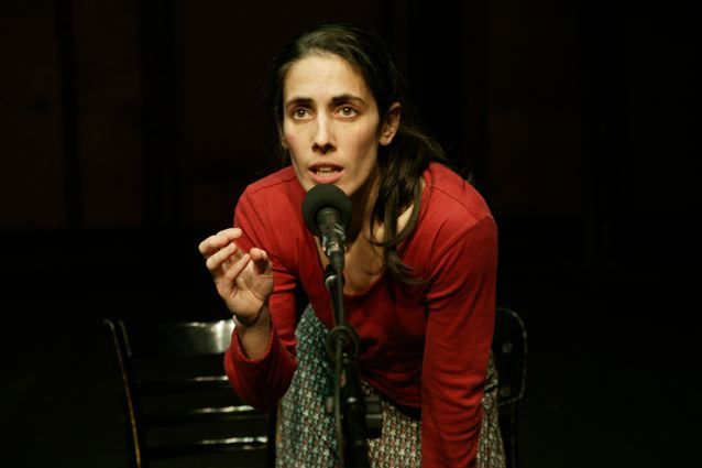 A woman in a red long sleeve t-shirt and printed pants bends over a microphone.
